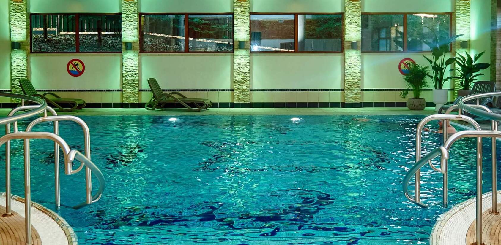 Kryty basen hotelowy z turkusową wodą, schodami i metalowymi poręczami, leżakami, roślinami i niebieskim oświetleniem