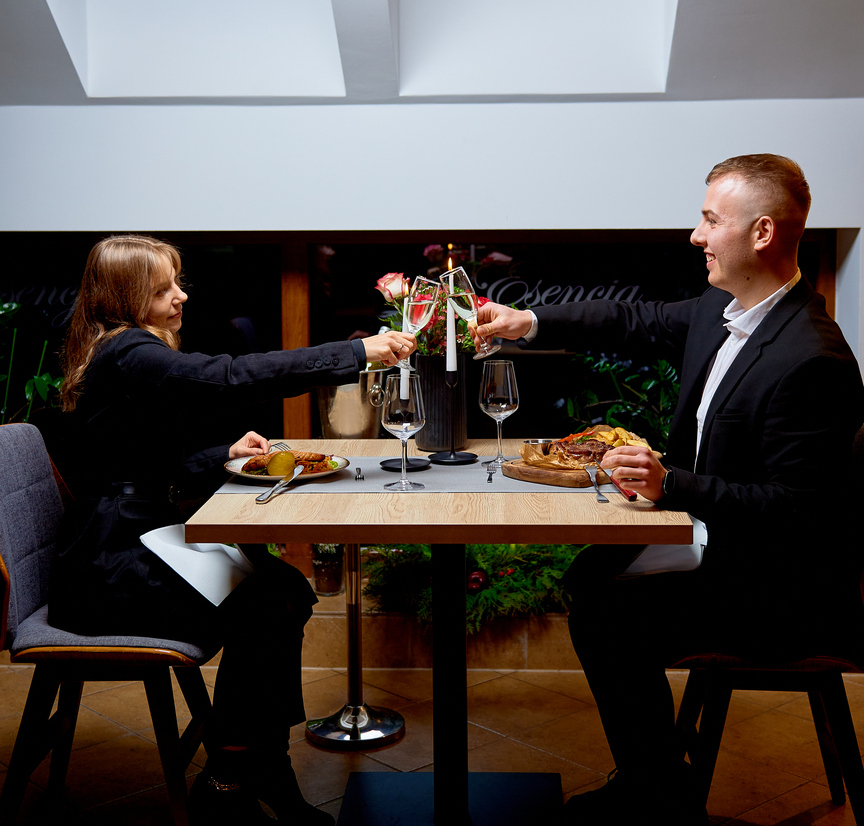 Couple in formal attire toasting champagne over a candlelit hotel dinner table with plated meals, roses centerpiece and skylight windows above