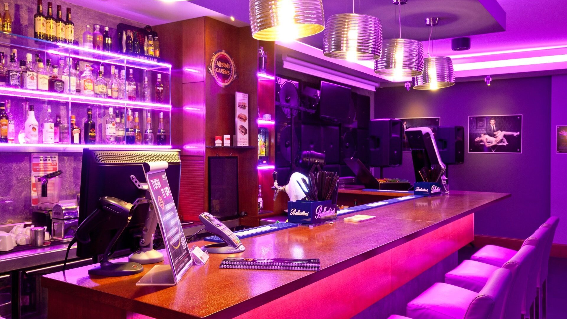 Nowoczesny bar hotelowy z fioletowym oświetleniem, wysokimi krzesłami i szerokim wyborem alkoholi na półkach za ladą.