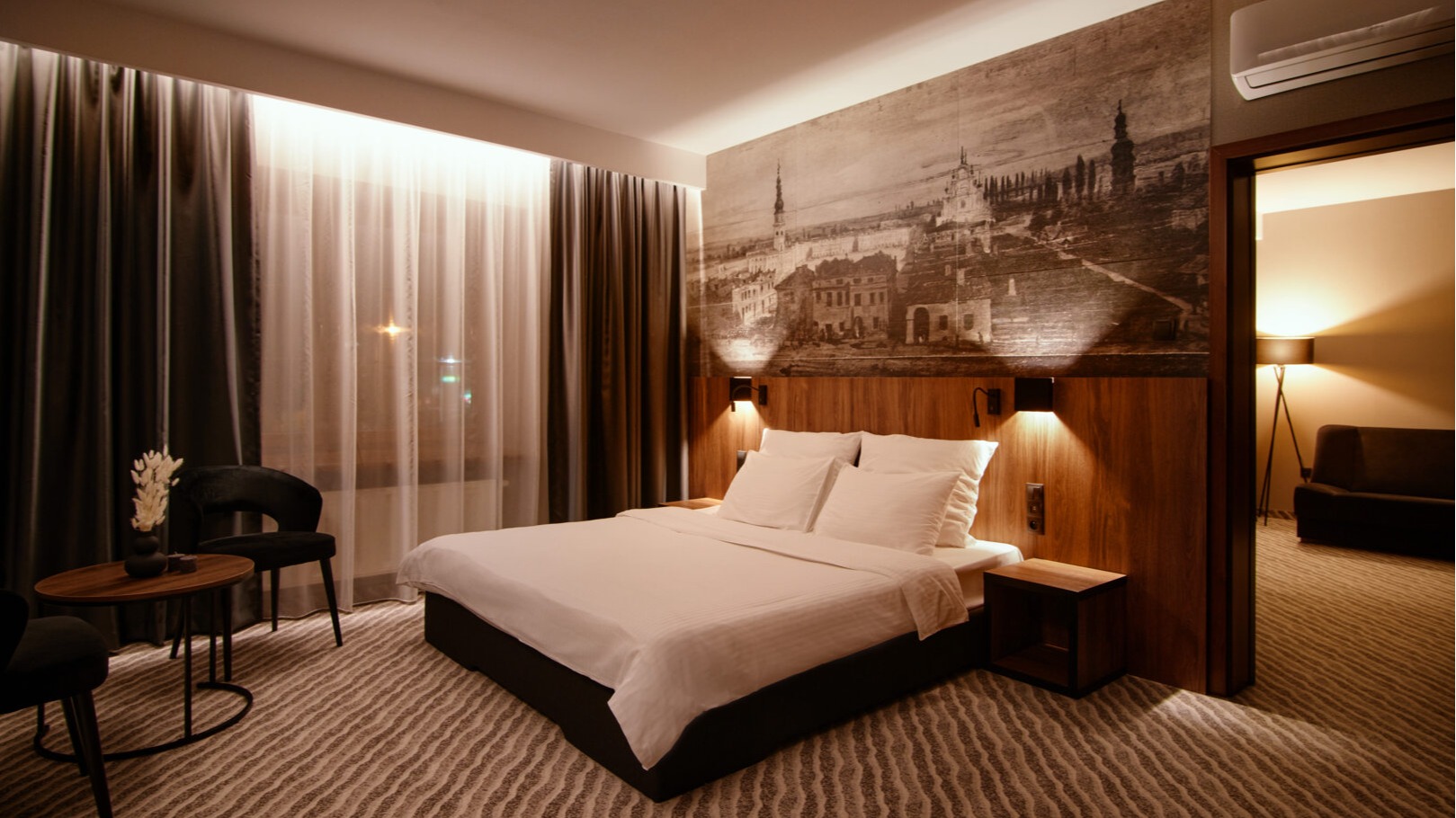 Elegancki pokój hotelowy z dużym łóżkiem, oświetleniem przy wezgłowiu i historyczną grafiką na ścianie.