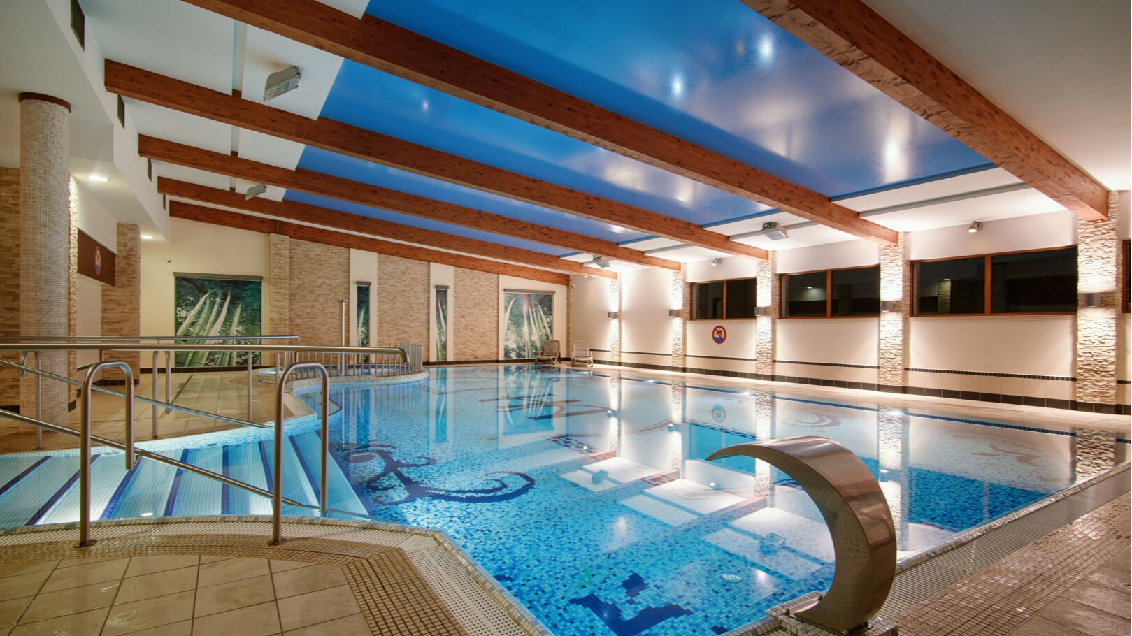Spacious indoor hotel pool with mosaic floor, wooden ceiling beams, and a relaxing, well-lit atmosphere.