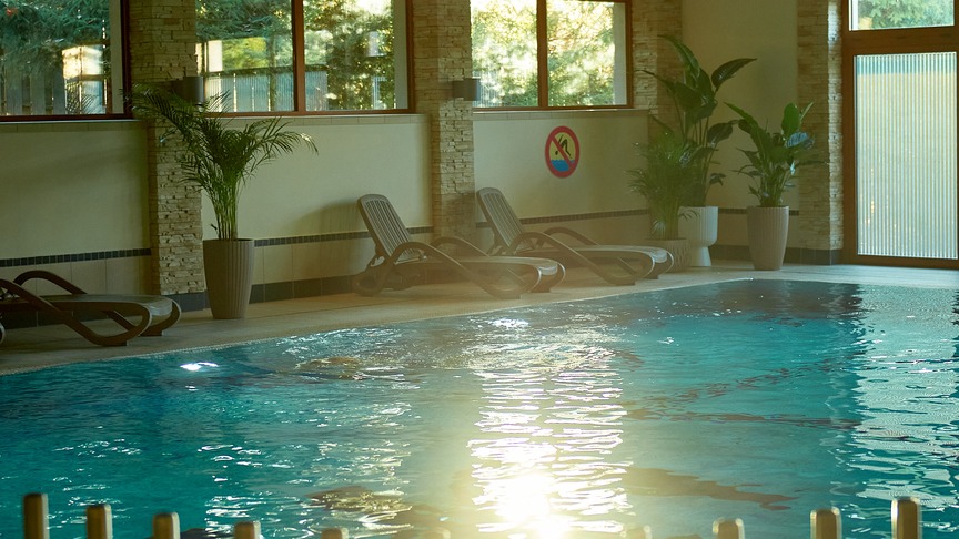Kryty basen hotelowy z turkusową wodą, drewnianymi belkami, leżakami i doniczkowymi palmami, relaksująca atmosfera