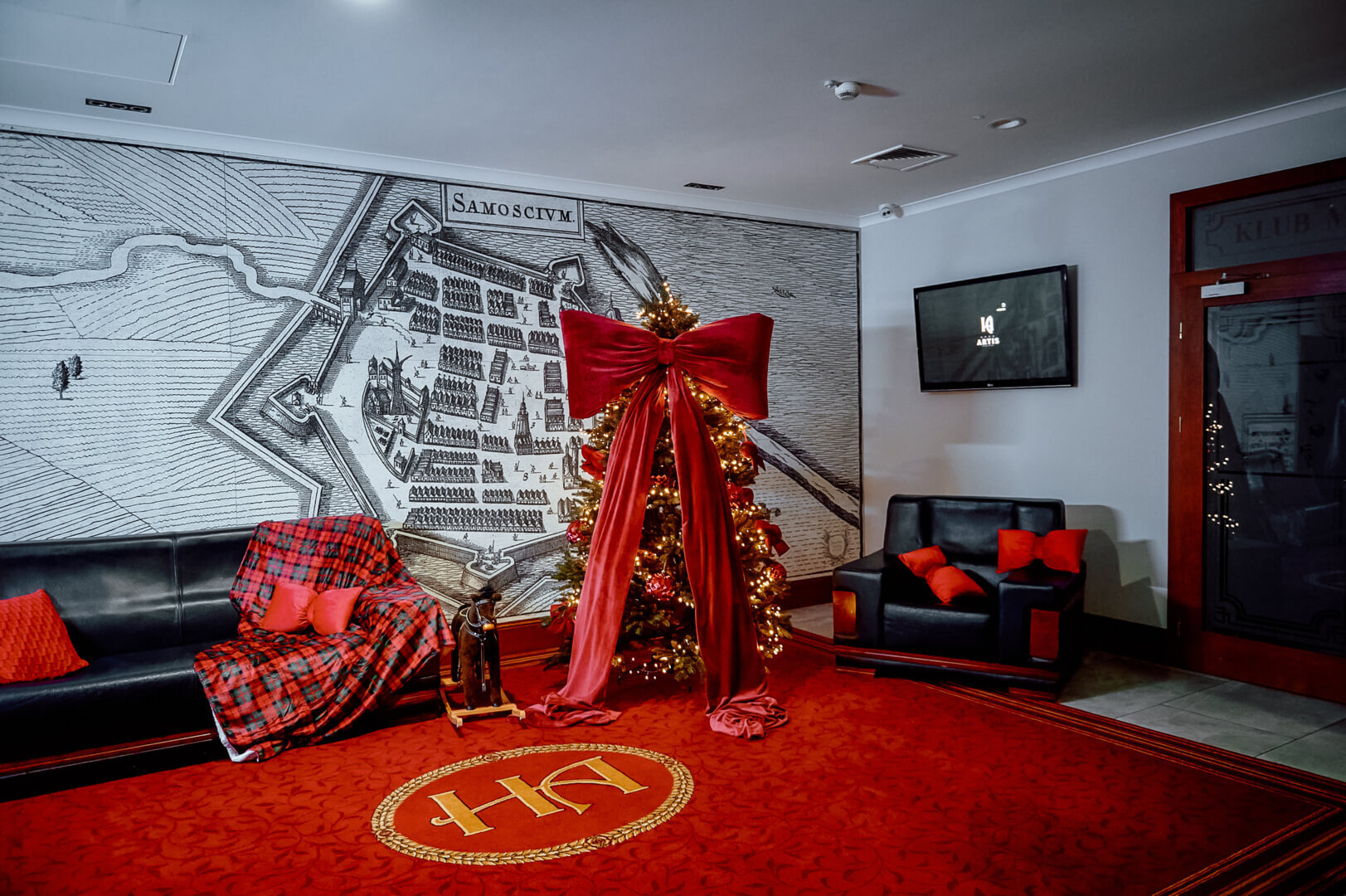 Hotel lounge with lit Christmas tree topped by oversized red velvet bow, black leather sofas with red cushions and an antique map mural.