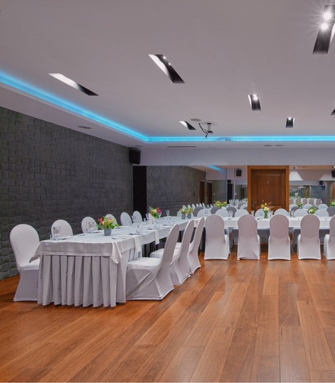 Elegancka sala konferencyjna z długimi stołami, białymi pokrowcami na krzesłach i dekoracjami kwiatowymi na parkiecie.