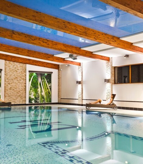 Stylowy, kryty basen hotelowy z mozaikowymi płytkami, dużymi oknami, drewnianymi belkami sufitowymi oraz leżakami z relaksującymi gośćmi w białych szlafrokach.