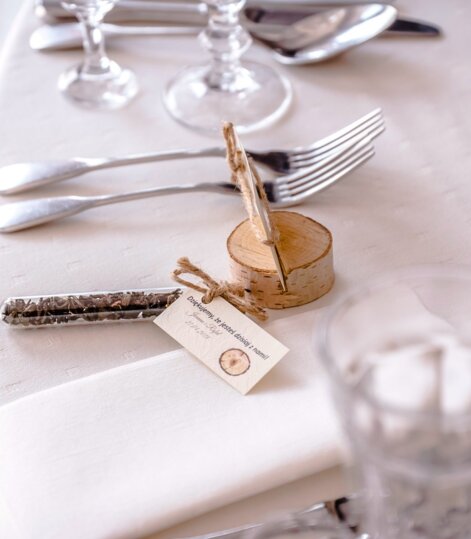 Elegancko nakryty stół z białym obrusem, rustykalną drewnianą podstawą na kartkę, sztućce i szklanką wody