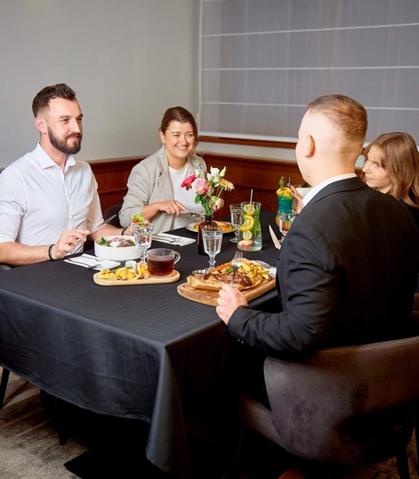 Four guests at a hotel restaurant table, enjoying plated meals, cocktails and a fresh floral centerpiece on a black tablecloth.