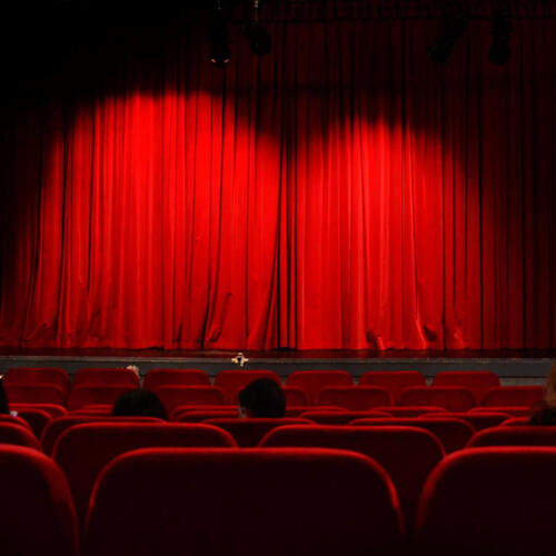 Kameralny teatr hotelowy z czerwoną aksamitną kurtyną, oświetloną sceną i wygodnymi czerwonymi fotelami, kilkoro widzów.
