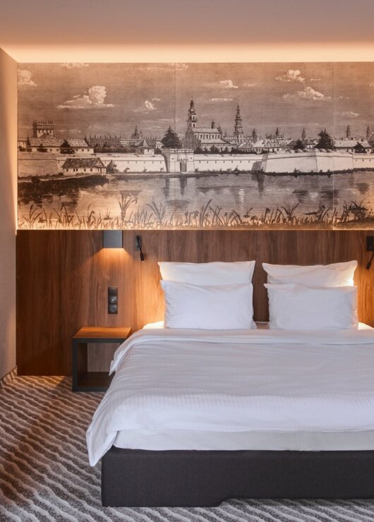 Nowoczesny pokój hotelowy z dużym łóżkiem, drewnianym wezgłowiem i panoramicznym muralem miasta nad wodą.