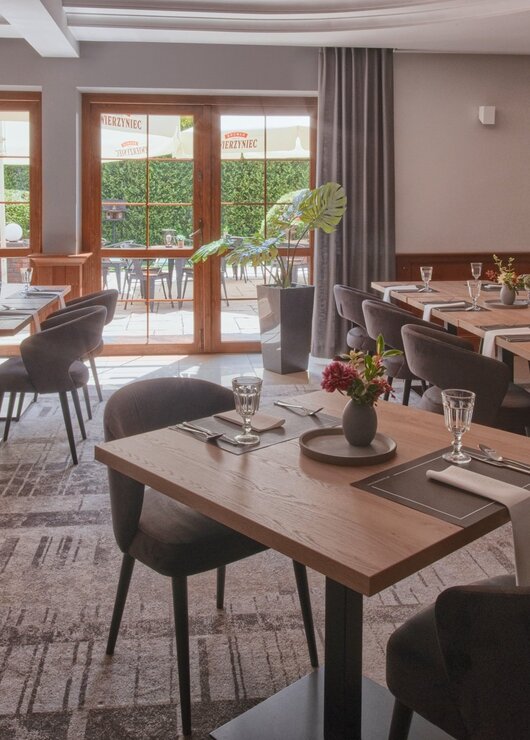 Cozy hotel dining area with wooden tables, comfortable grey chairs, and access to an outdoor terrace with greenery.
