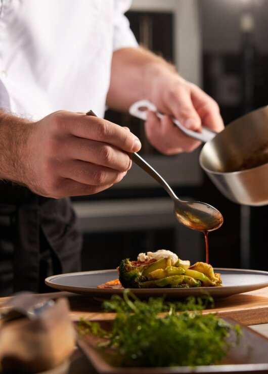 Szef kuchni w białej koszuli przygotowuje danie, polewając sosem warzywa na talerzu, z drewnianą deską i ziołami obok.