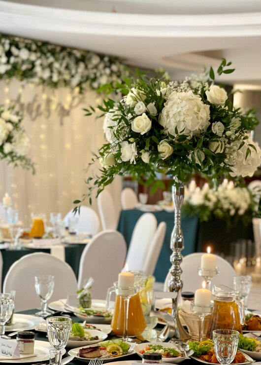 Elegancka sala weselna z wysokimi bukietami białych kwiatów, świecami i stołami zastawionymi świeżymi potrawami.