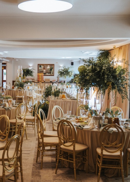 Elegancka sala bankietowa z okrągłymi stołami, złotymi krzesłami i bujnymi zielonymi dekoracjami na wesele lub uroczystość.