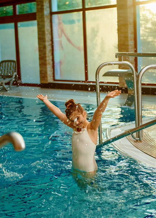 Young girl playing and splashing in a sunlit indoor hotel pool by stainless handrails and deck loungers.