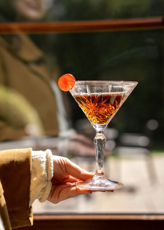 Crystal martini glass with amber cocktail and maraschino cherry, held by a guest near a sunlit hotel bar window.