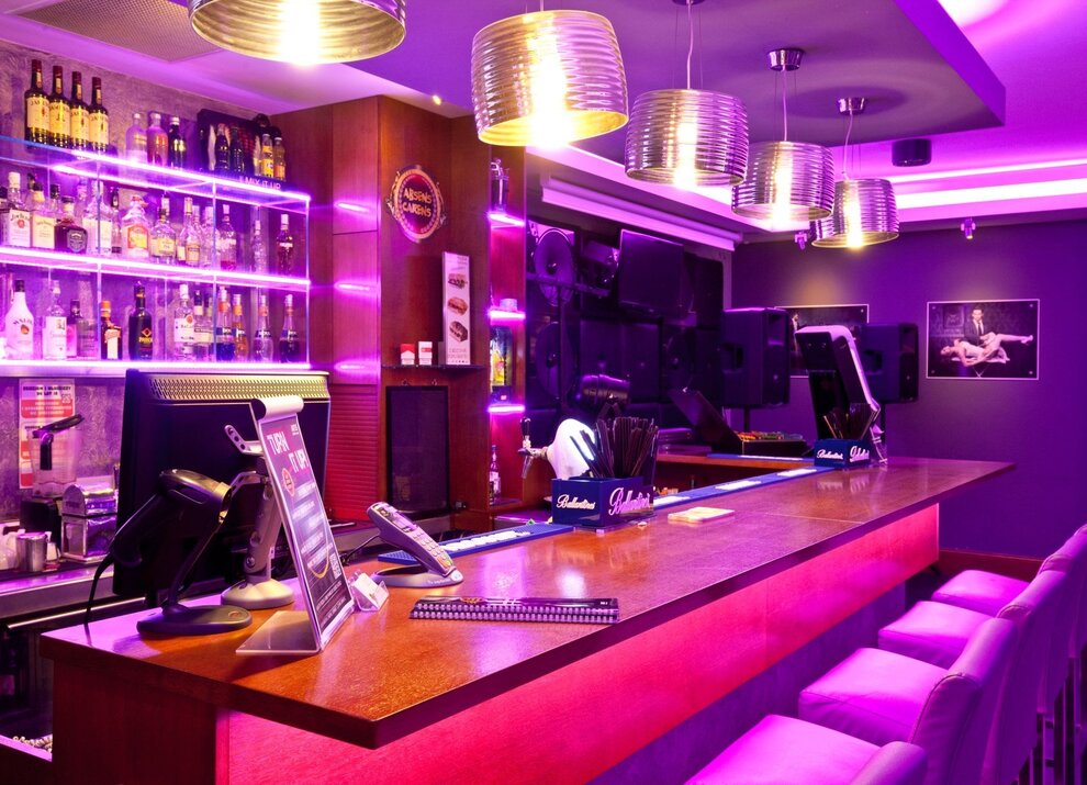 Nowoczesny bar hotelowy z fioletowym oświetleniem, wysokimi krzesłami i szerokim wyborem alkoholi na półkach za ladą.