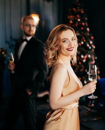 Elegant woman in satin dress holding champagne glass in festive lounge with man in tuxedo and decorated Christmas tree.