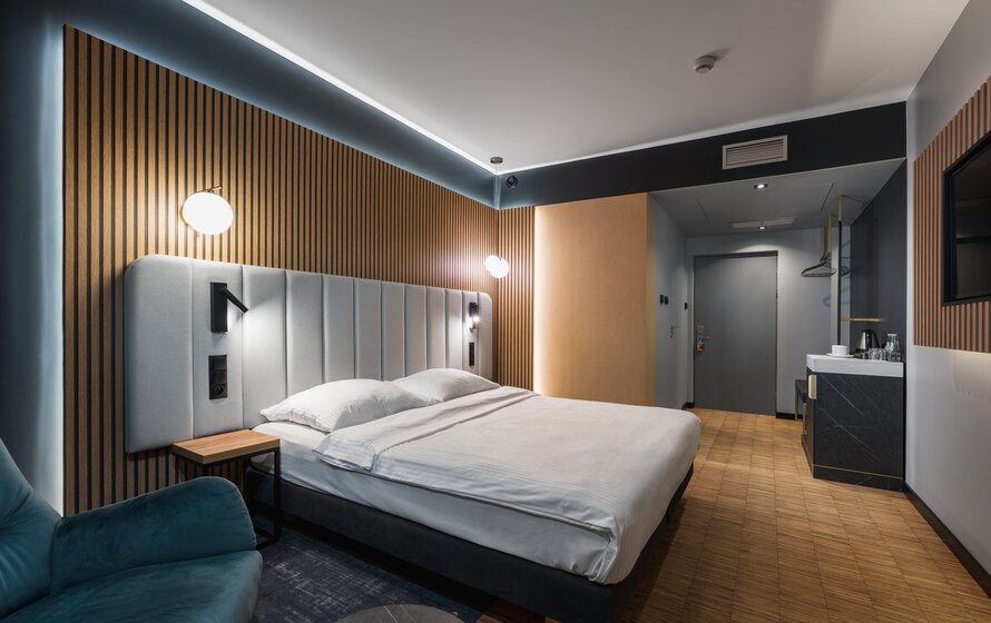 Modern hotel room with large bed, soft upholstered headboard, LED lighting, and a comfortable armchair near the bed.
