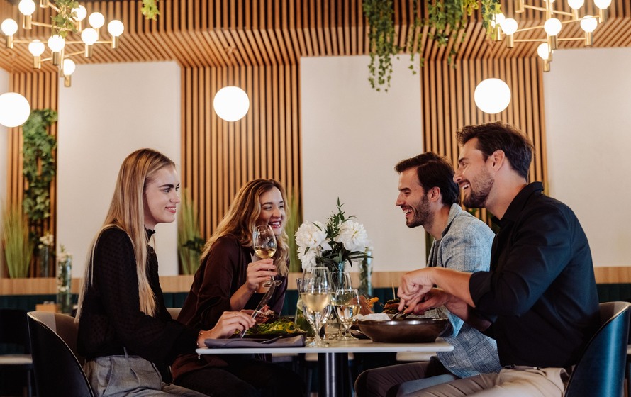 Four guests enjoying food and wine in a modern restaurant with wooden wall panels and floral centerpiece.