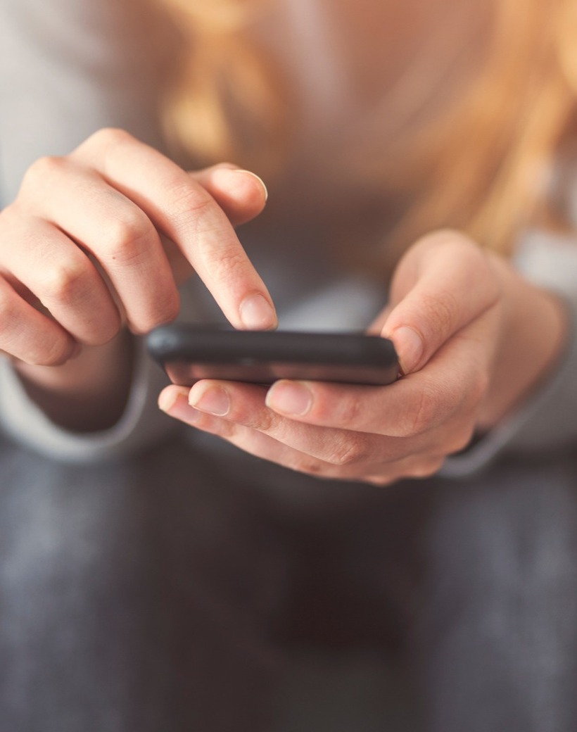 Close-up of hands tapping a smartphone screen, seated person with blurred blonde hair in the background