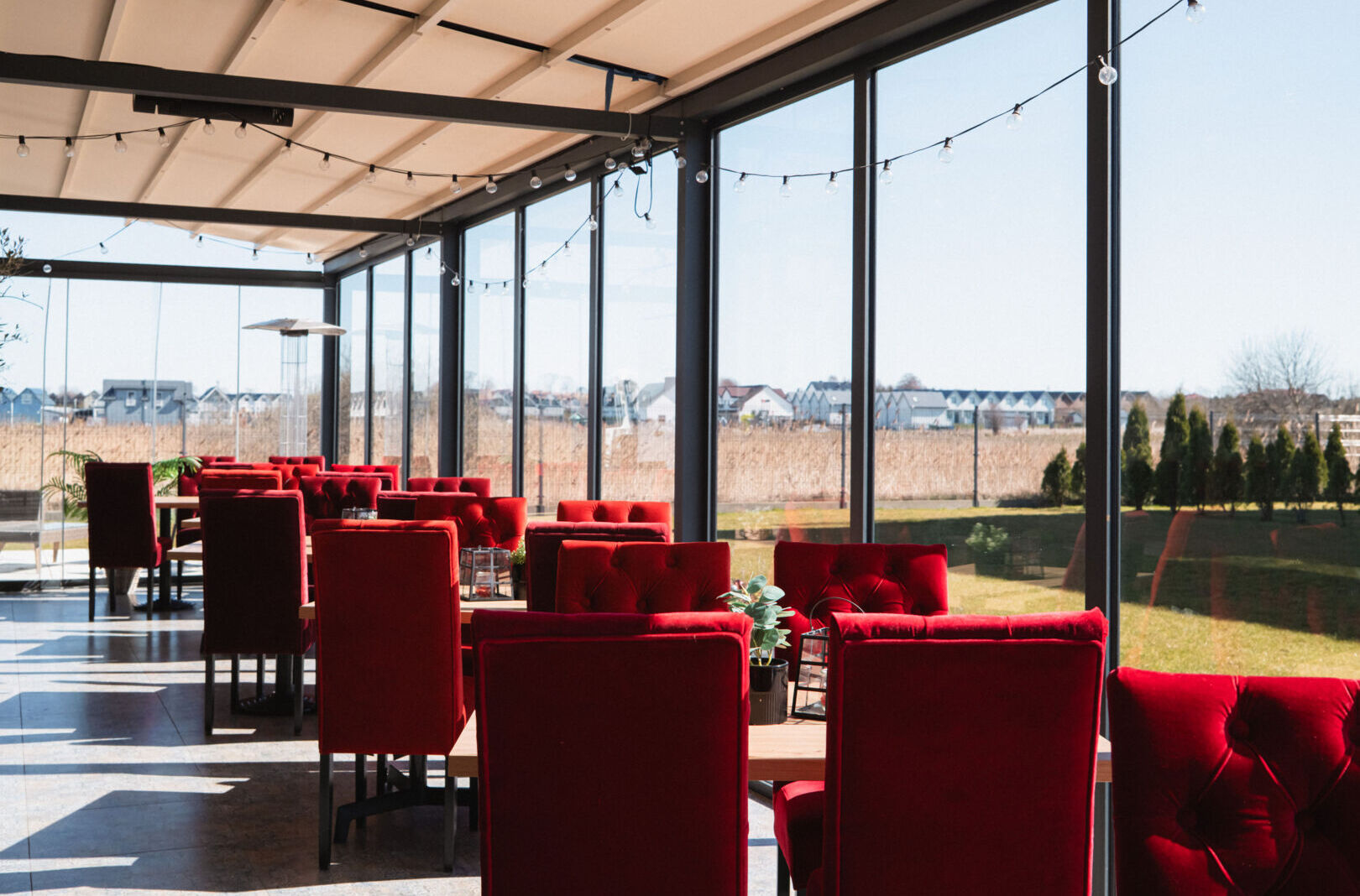 Jasna sala restauracyjna z czerwonymi, pikowanymi krzesłami i dużymi oknami z widokiem na ogród i okoliczne domy.