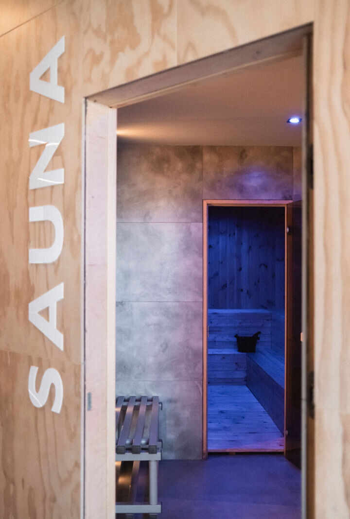 Drzwi do sauny z drewnianą ławką na zewnątrz i wnętrzem z ciemnego drewna oraz wiadrem do wody.