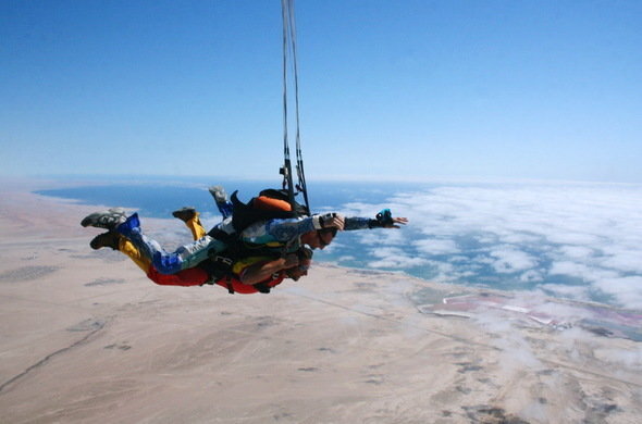 Tandem skydive above arid coastline, instructor and guest in harness over desert and clouds, panoramic aerial view