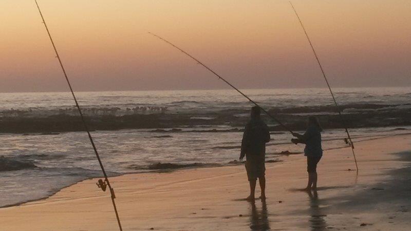Two people surf-fishing at sunset on a sandy beachfront, rods planted in wet sand beside gentle waves