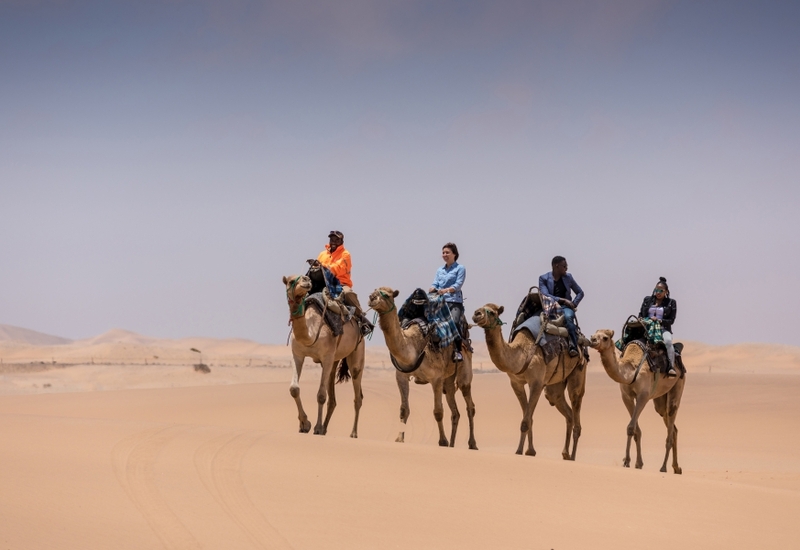Guided camel ride with four riders crossing golden desert dunes under a pale blue sky