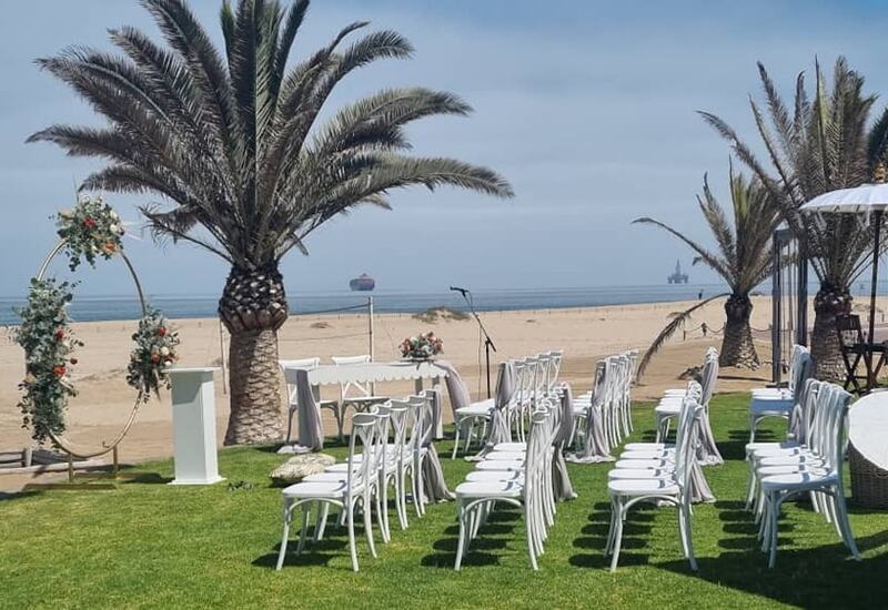 Beachfront wedding setup on grassy lawn with white chairs, floral arch, palm trees and ocean view for seaside ceremony.