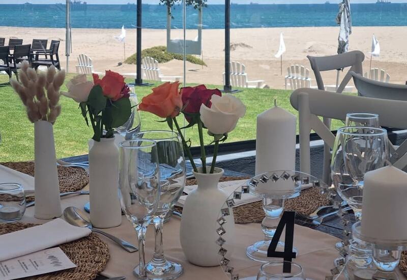 Beachfront dining table set with roses, candles, wine glasses and woven placemats, glass wall opening to sandy beach and ocean.