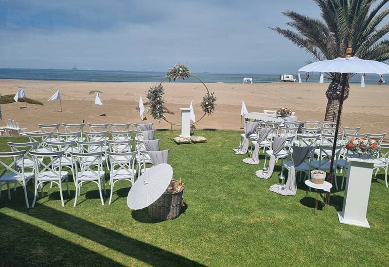 Beachfront wedding setup on green lawn with white chairs, floral hoop arch facing ocean, parasols and palm tree.