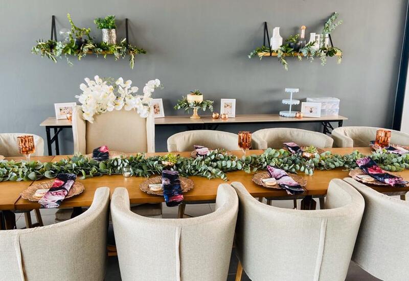 Long wooden dining table with beige upholstered chairs, eucalyptus garland, floral napkins, rose-gold goblets and orchid.