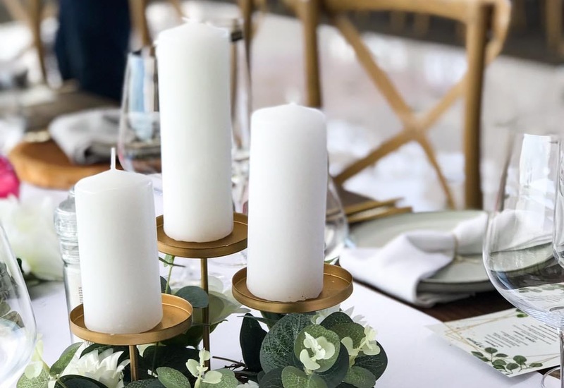 Elegant table centerpiece - three white pillar candles on a gold stand with eucalyptus and a white rose, wine glasses.