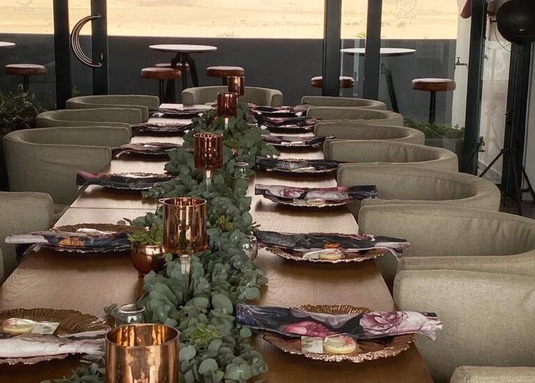 Banquet table set for private dinner with eucalyptus garland, rose‑gold goblets, floral napkins and cushioned chairs overlooking desert dunes