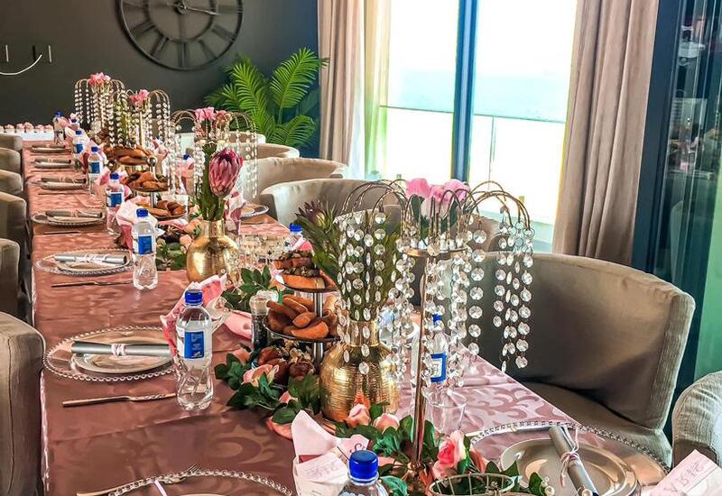Elegant long dining table set for event with pink linens, crystal candelabras, charger plates, bottled water, plush chairs by sunlit floor-to-ceiling windows