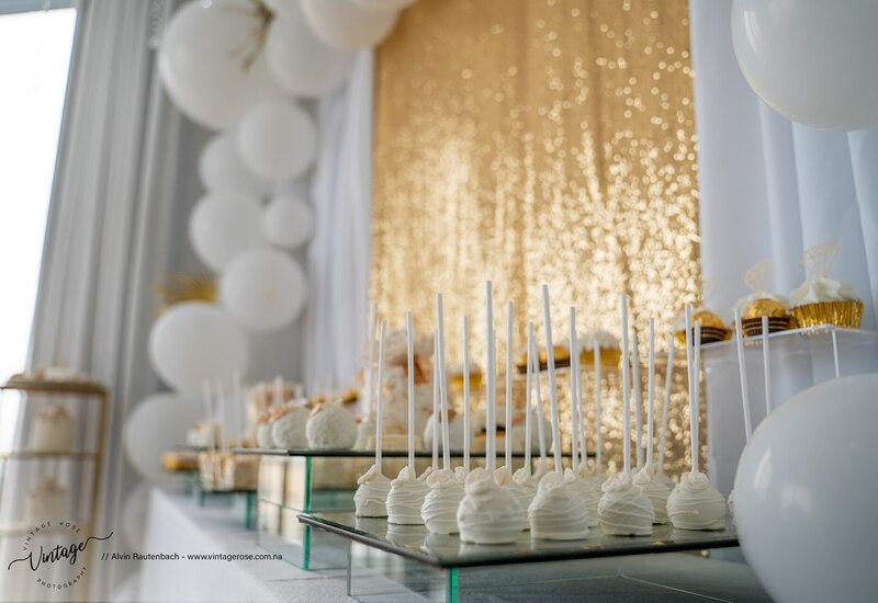 Elegant white-and-gold dessert table with cake pops, cupcakes and white balloons against a sparkling gold sequin backdrop