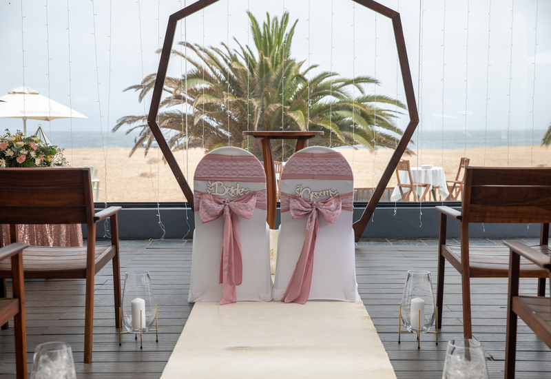 Beachfront wedding setup with Bride and Groom chairs draped in pink bows beneath a wooden hex arch, ocean view.