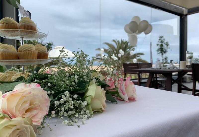 Pastel rose and baby's-breath centerpiece on a white table with tiered cupcakes, balloons and dining tables on a glass-enclosed seaside terrace.