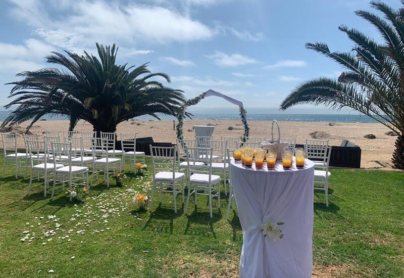 Beachfront wedding setup with white chairs and floral arch on green lawn, palm trees and ocean view, cocktail table with juices.