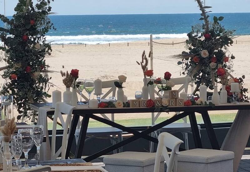 Beachfront wedding sweetheart table beneath a floral hoop arch with candles, red and white roses and ocean waves beyond.