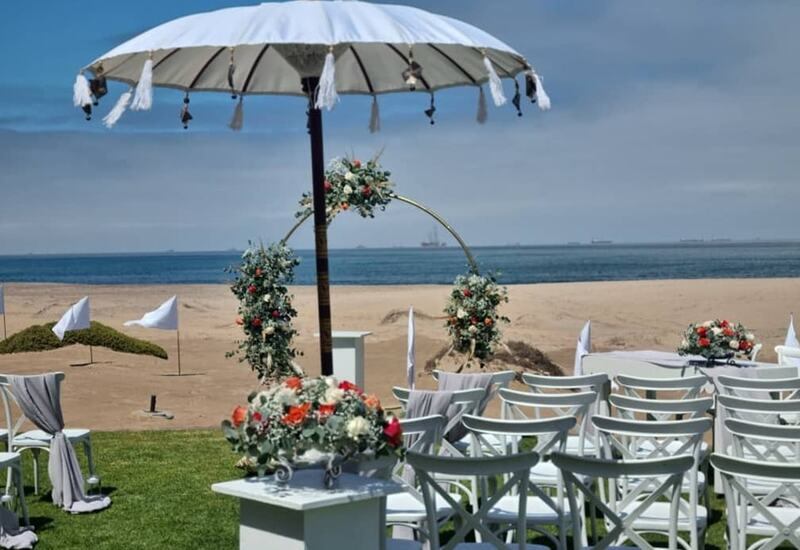 Beachfront wedding setup with white chairs, floral arch and decorative parasol facing calm ocean and sandy shore