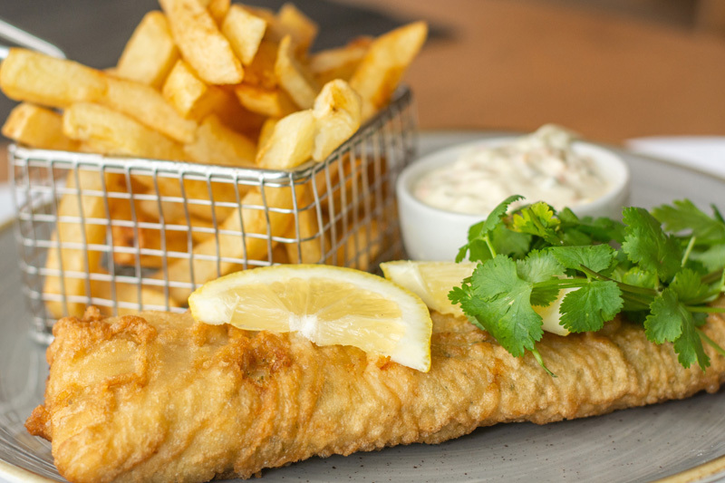 Beer-battered fish fillet with lemon wedges, fresh cilantro, tartar sauce and thick-cut fries served in a wire basket