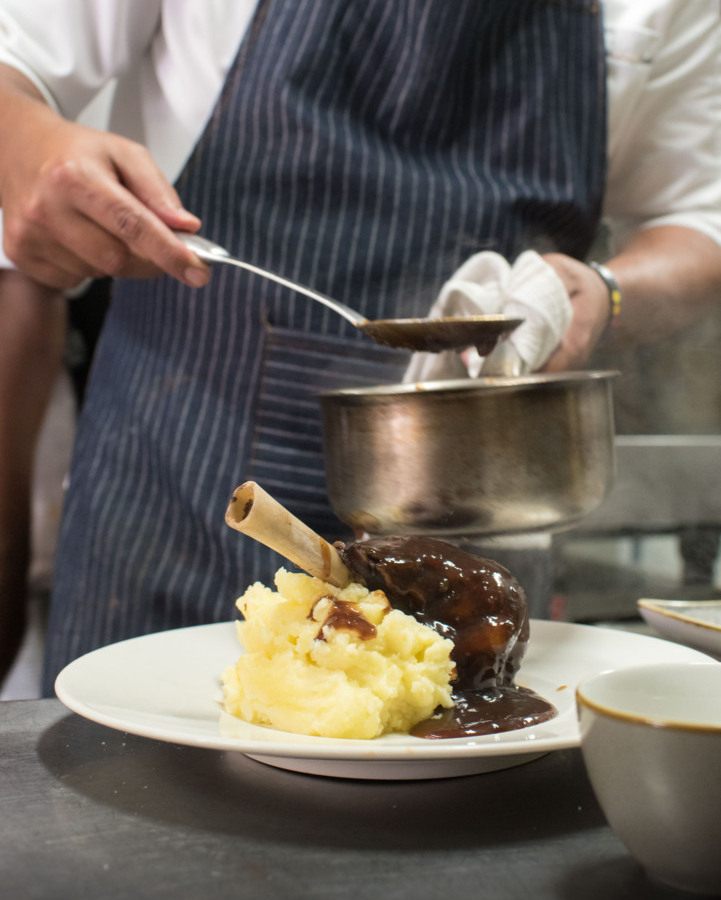 Chef in pinstripe apron ladles rich gravy over a braised lamb shank served with creamy mashed potatoes in restaurant kitchen