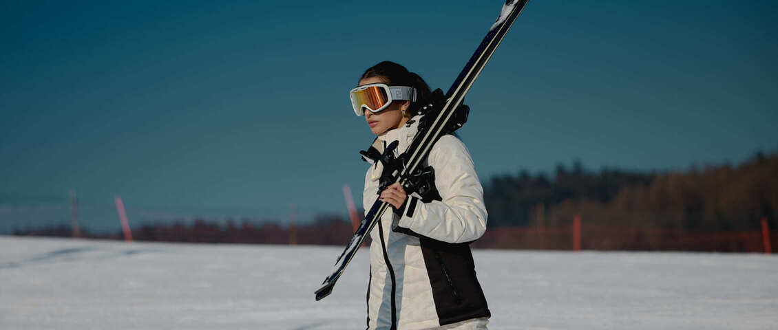 Skier in white jacket and goggles carrying skis over shoulder across a snowy slope under clear blue sky