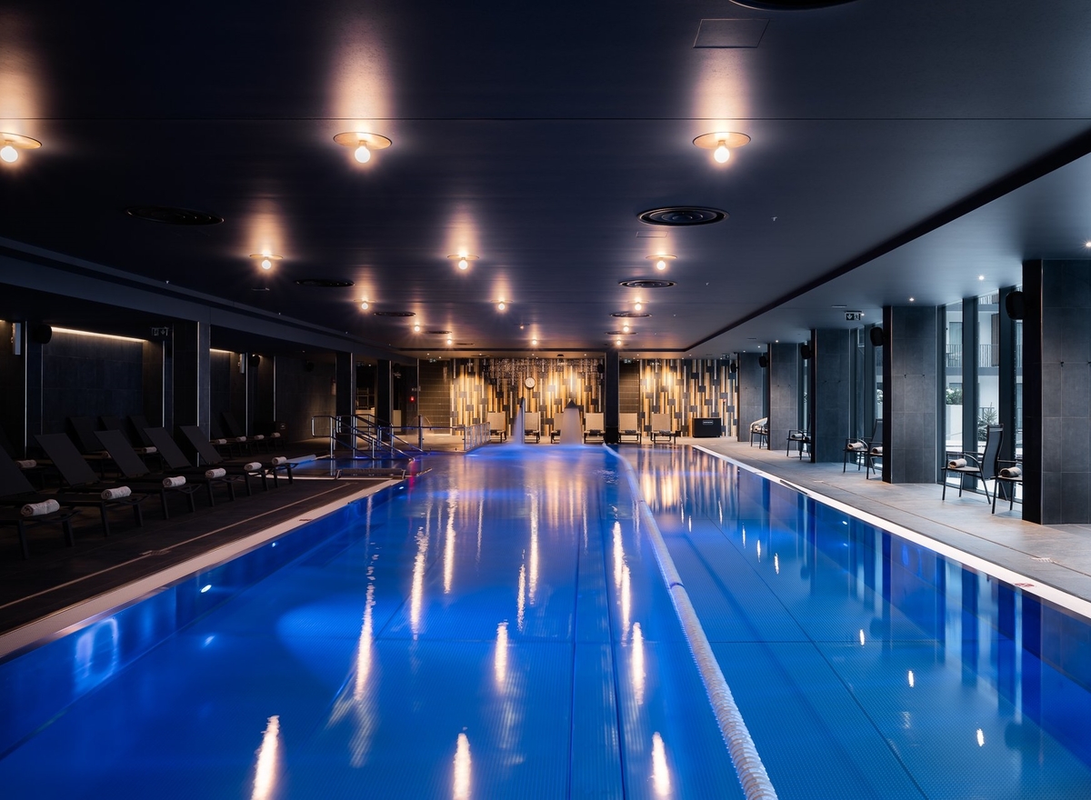 Kryty basen hotelowy z niebieską wodą, torami pływackimi, leżakami, ciepłym oświetleniem i panoramicznymi oknami.