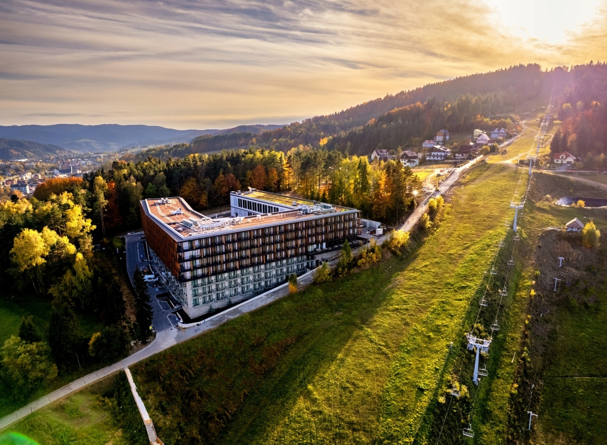 Modern multi-story hotel with balconies beside a ski slope and chairlift, autumn forest and panoramic mountain valley at sunset.