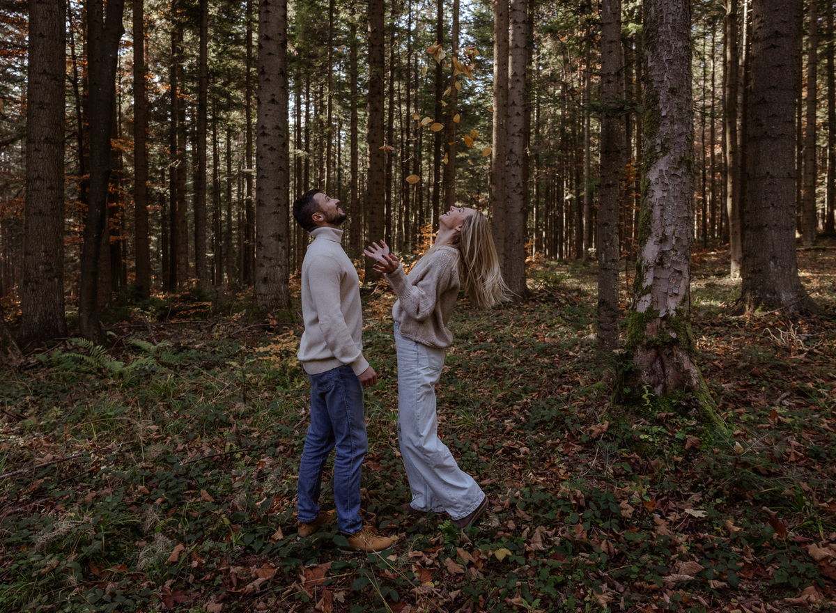 Couple in cozy sweaters tossing autumn leaves in a tall pine forest, playful outdoor moment