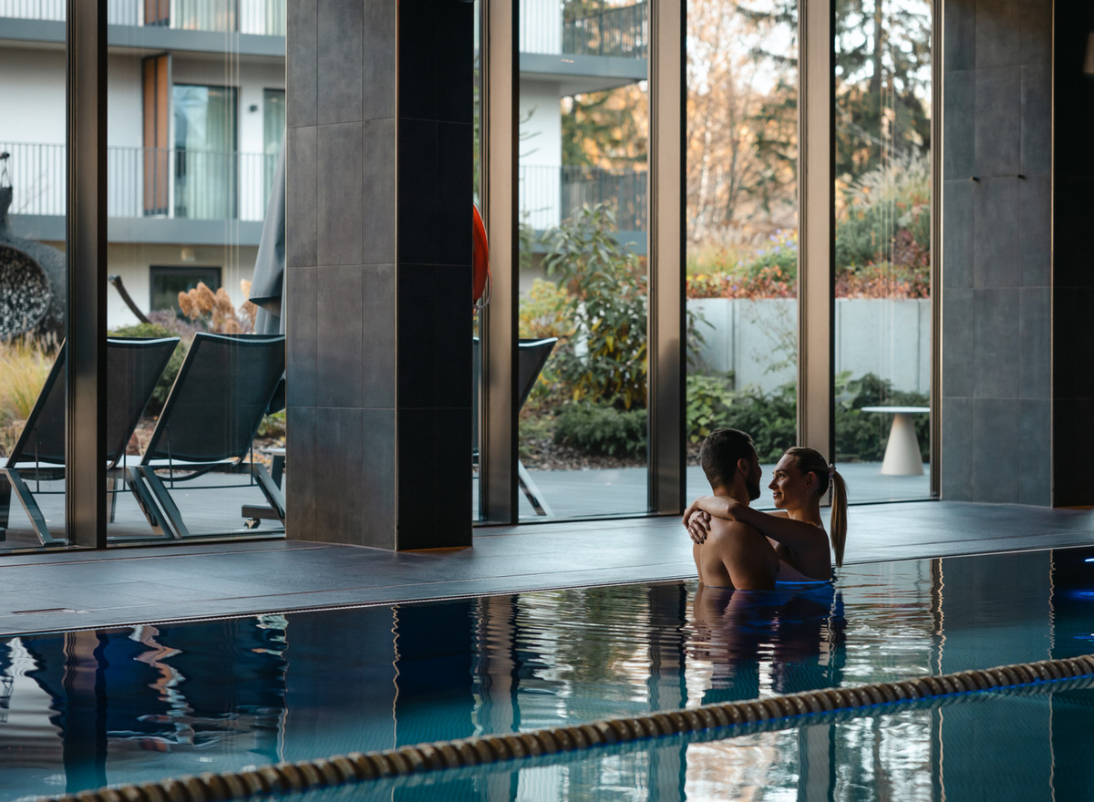 Couple embracing in a hotel indoor pool by floor-to-ceiling windows, loungers outside, modern spa-like atmosphere