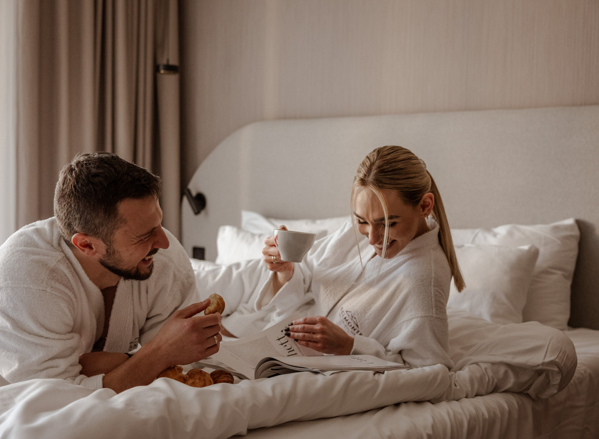 Couple in plush robes enjoying breakfast in bed, coffee, croissants and a magazine in a cozy hotel room.